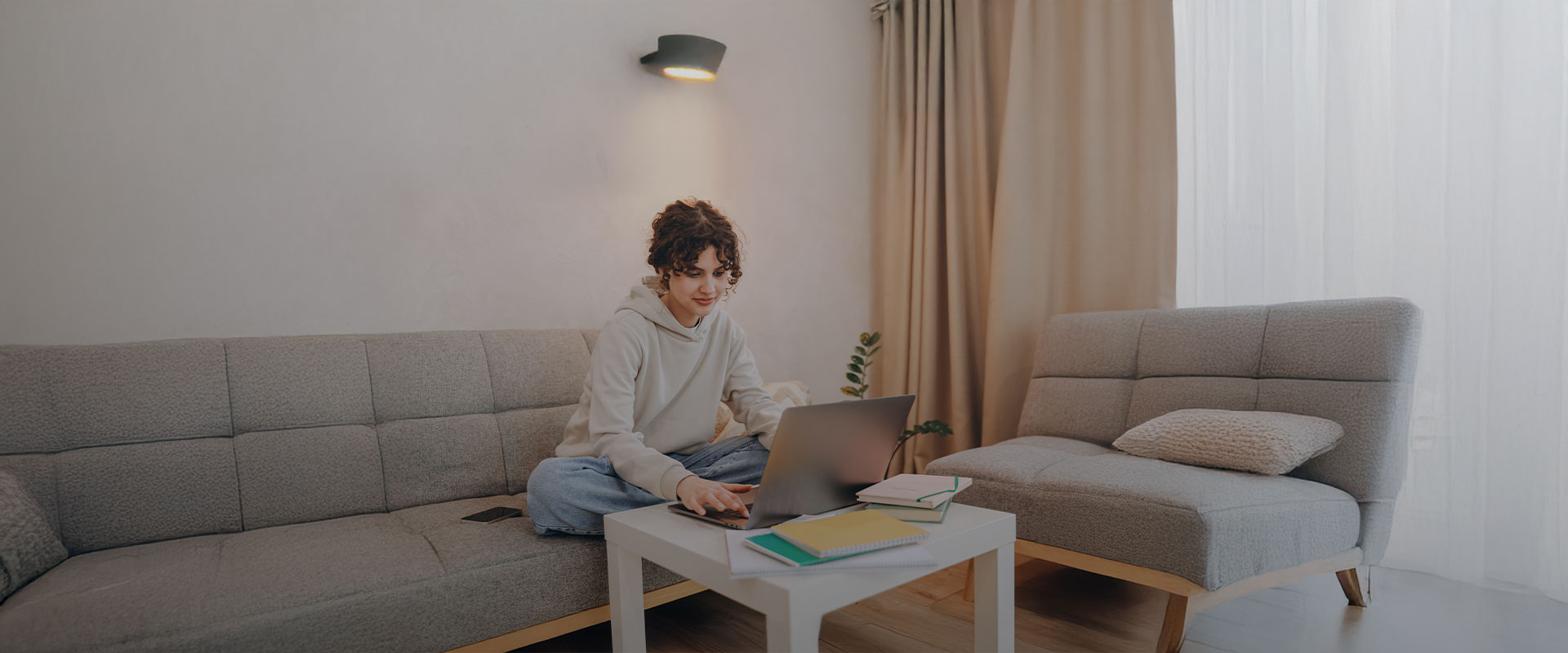 Persona joven sentada en un sofá usando un notebook sobre una mesa de centro, en un ambiente hogareño con cuadernos y cortinas al fondo.